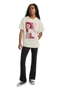 Cream-colored T-shirt with bold red and pink Converse graphic design, short sleeves, relaxed fit, paired with black pants and sneakers.