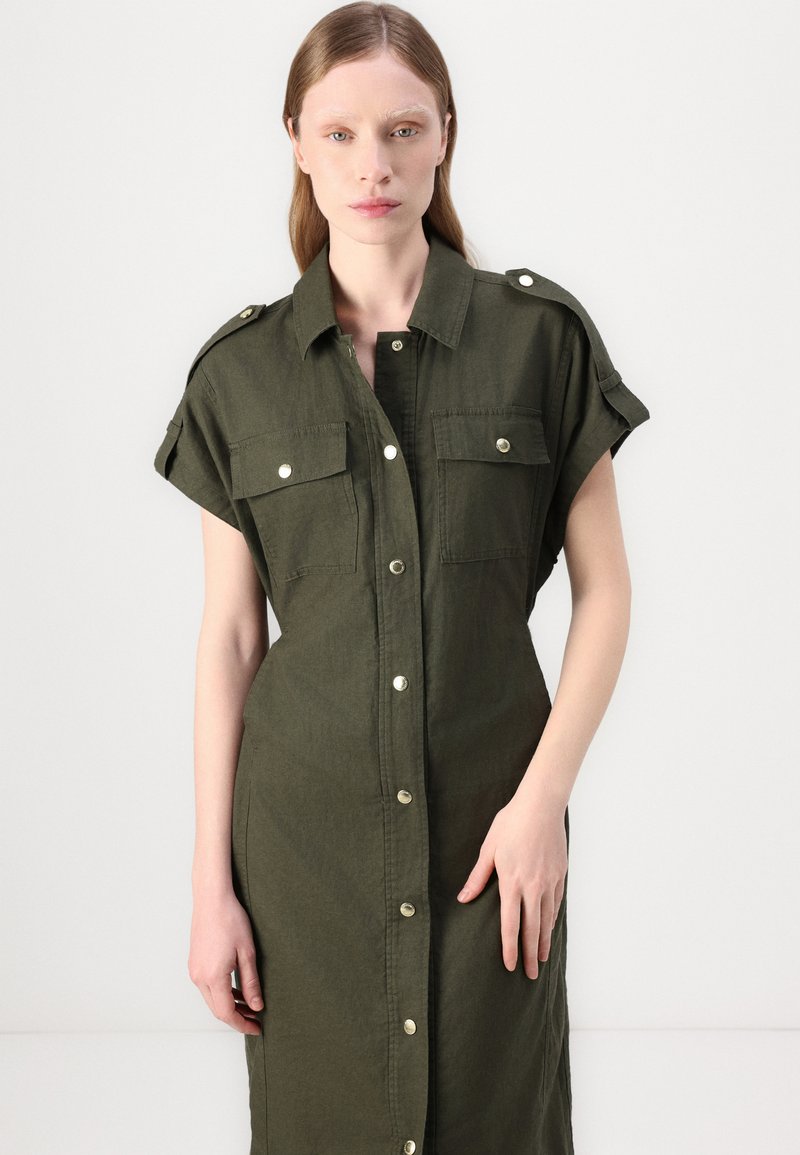 Zielona lniana sukienka koszulowa z krótkimi rękawami, epauletami na ramionach, dwiema kieszeniami na piersi oraz z przodem zapinanym na guziki. Gładka tekstura, do kolan.