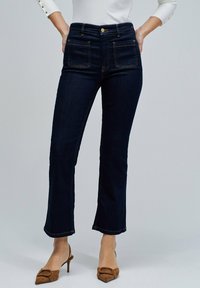 Mujer que lleva unos jeans rectos de tiro alto en azul oscuro con bolsillos frontales y unos zapatos de tacón marrones con punta afilada.