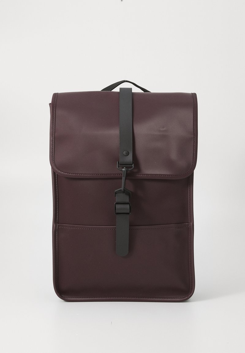 Mochila color burdeos con cierre de solapa y correa negra, que cuenta con un bolsillo frontal y un diseño minimalista sobre un fondo blanco liso.