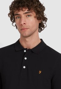 Zwarte polo shirt met een drieknoopsluiting, witte knopen, korte mouwen en een klein oranje geborduurd logo op de linkerkant van de borst.