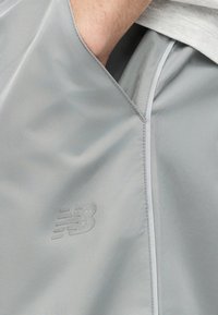 Χέρι που γλιστράει στην πλαϊνή τσέπη αθλητικών σορτς New Balance ανοιχτού γκρι χρώματος με εμφανές λογότυπο και τελείωμα ελαφριάς χρωματιστής μπλούζας.