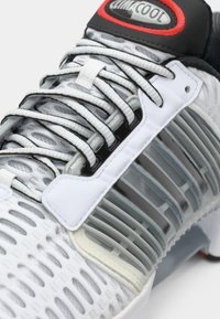 Witte sportschoen met een geprofileerde mesh bovenwerk, grijze rubberen accenten, zwart en rode branding, en een uniek ventilatieontwerp.