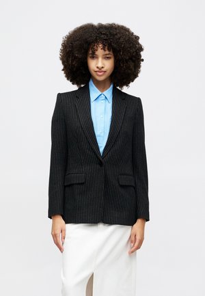 Sort stribet blazer med nyklippede revers, to frontlommer og lukning med en enkelt knap, båret over en lys blå skjorte.