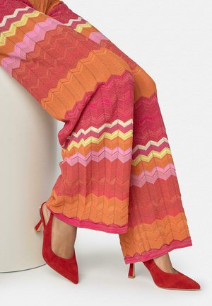 Benen met wijde broekspijpen in oranje, roze en gele chevronstrepen, gecombineerd met rode suède puntige hoge hakken.