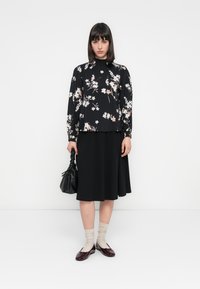 Blusa nera a maniche lunghe con motivo floreale, abbinata a una gonna nera a trapezio. Gli accessori includono una borsa nera texturizzata e scarpe bordeaux con calzini.