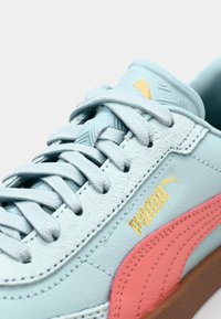 Baskets bleu clair en cuir texturé, avec accent rose sur le côté, logo doré et lacets plats gris. Semelle extérieure en caoutchouc, forme arrondie.