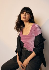 Roze geribd topje met een vierkante halslijn, gecombineerd met een zwarte blazer. Model zit op een kruk, draagt donkere spijkerbroek en een riem. Zachte textuur.