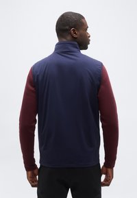 Άνδρας με την πλάτη γυρισμένη, φοράει μπλε σκούρο γιλέκο χωρίς μανίκια πάνω από πουκάμισο με μακριά μανίκια σε μπορντό χρώμα και μαύρο παντελόνι, μπροστά σε απλό ανοιχτό φόντο.