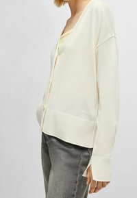 Krämfärgad kofta i mjukt tyg, med V-ringning, knappar, ribbad mudd och en diskret "BOSS"-logotyp vid kanten. Bärs över en ljus topp.