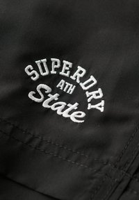 Zwarte stof met witte geborduurde tekst "SUPERDRY ATH State." Heeft een zachte textuur en een gebogen ontwerp in de letters.