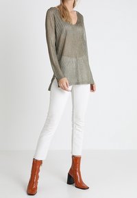 Olivově zelený svetr s dlouhým rukávem a výstřihem do V s volným pleteným vzorem; doplněný bílými přiléhavými kalhotami a lesklými kotníkovými botami v barvě spálené oranžové.