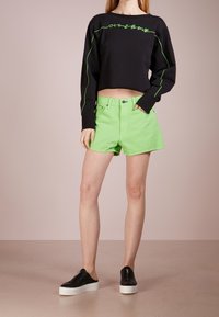 Sweat-shirt court noir avec des coutures et des lettres en vert, associé à un short vert vif et des chaussures noires à enfiler avec des semelles blanches.