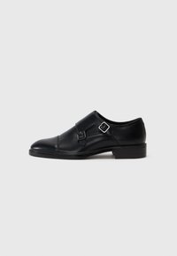 Black leather shoe with a rounded toe, featuring two silver buckles and minimal detailing. Low heel and smooth texture.