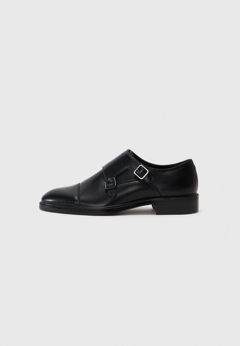 Black leather shoe with a rounded toe, featuring two silver buckles and minimal detailing. Low heel and smooth texture.