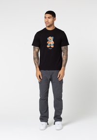 Black cotton t-shirt featuring a colorful bear graphic on the front, paired with gray cargo pants and white sneakers. Smooth texture.
