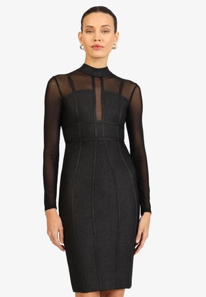 Frau trägt ein eng anliegendes schwarzes Kleid mit transparenten langen Ärmeln und obere Brustpartie, stehend vor einem schlichten weißen Hintergrund.