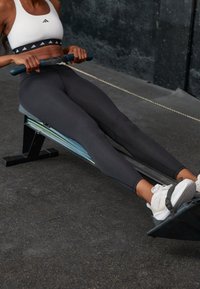 Máquina de remo con un elegante marco negro, detalles en azul y un asiento texturizado. El usuario lleva pantalones de chándal grises y zapatos blancos, mientras sostiene un mango azul.