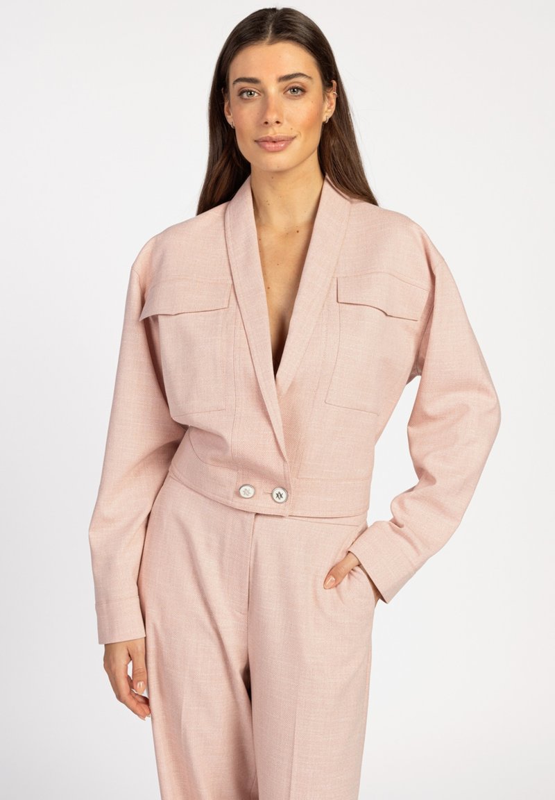 Lichtroze op maat gemaakte jumpsuit met lange mouwen, een shawl-kraag en twee borstzakken. Heeft wijde pijpen en een dubbele knoopsluiting in de taille.