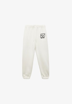 Białe dresowe spodnie z elastycznymi mankietami i paskiem w talii, z dodatkowym sznurkiem i nadrukiem "25 New York" w czerni na górnej lewej stronie uda.