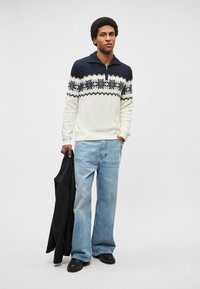 Biały sweter dziany z ciemnoniebieskimi akcentami i geometrycznym wzorem, w połączeniu z jasnoniebieskimi szerokimi spodniami dżinsowymi i czarnymi butami, trzymający czarną kurtkę.