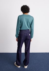 Mörkblå denimbyxor ihop med en långärmad turquoise skjorta. Byxorna har synliga bakfickor och gula sömmar. Skorna är ljusa.