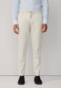 Helle beige Hose mit einem Kordelzugbund, glatter Textur, zulaufenden Beinen und subtilem Glanz, kombiniert mit dunklen Schuhen.