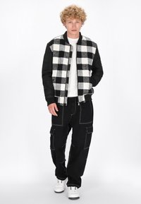 Czarna i biała kurtka bomber z wzorem w kratę oraz kontrastowymi czarnymi rękawami, zestawiona z czarnymi spodniami cargo z białym przeszyciem, noszona z białymi trampkami.