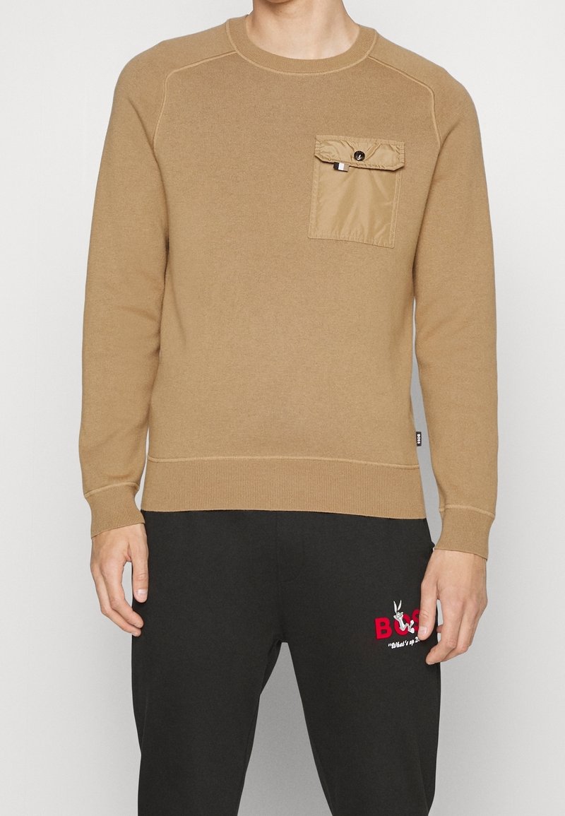 Brauner Pullover mit geripptem Rundhalsausschnitt, langen Ärmeln und einer Brusttasche aus kontrastierendem, sandfarbenem Material. Getragen mit schwarzen Hosen, die ein Logo tragen.
