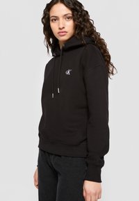 Μαύρο φούτερ φτιαγμένο από βαμβακερό μείγμα, με μπροστινή τσέπη, ρυθμιζόμενο κορδόνι και κεντημένο λογότυπο στο στήθος.