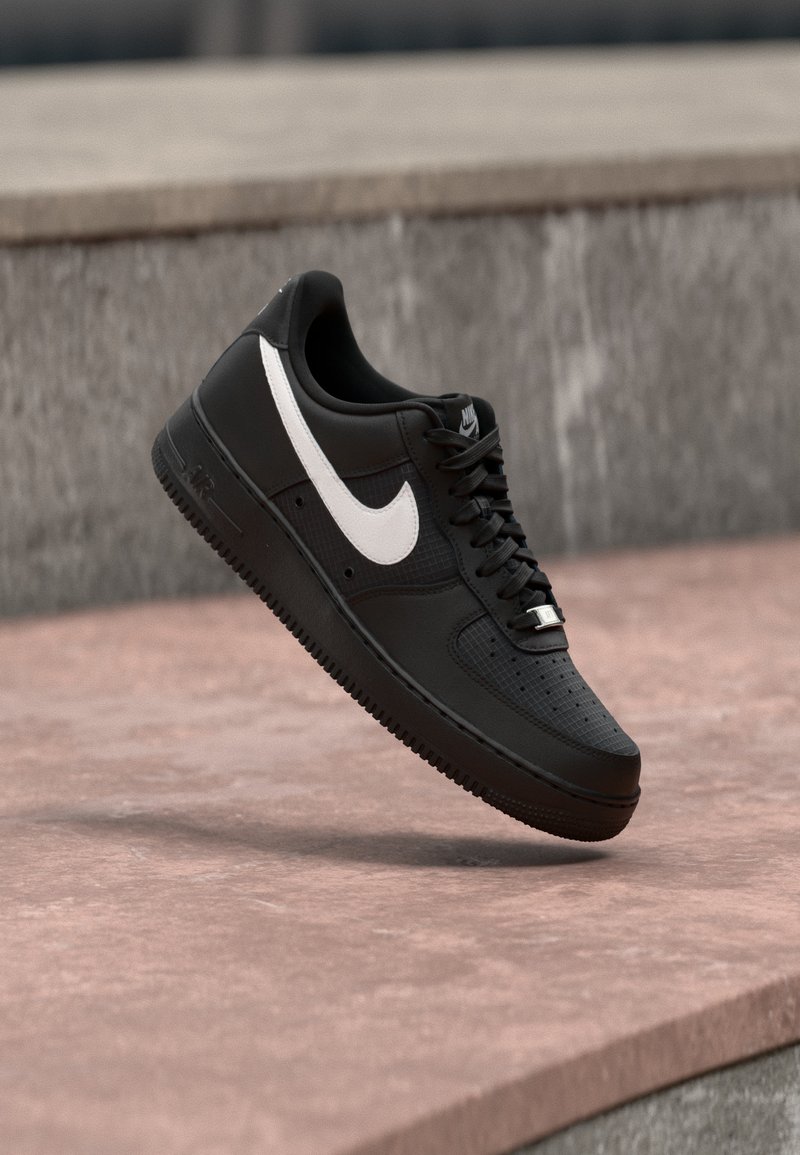 Pantof Nike Air Force 1 negru cu logo alb Swoosh plutind deasupra unei suprafețe de piatră și cu un perete texturat gri în fundal.