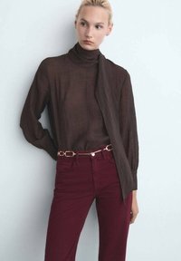 Femme portant un blouse transparente marron foncé à col montant avec un long nœud et un pantalon taille haute bordeaux avec une ceinture en chaîne dorée.