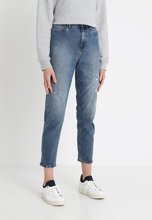 Jean en denim bleu avec une coupe droite, présentant des détails déchirés. Associé à des baskets blanches et un sweat-shirt gris.