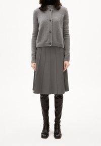 Kvinna iklädd en grå knappad cardigan, veckad grå kjol och svarta knähöga läderstövlar, stående mot en vit bakgrund.