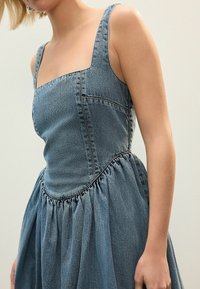 Denimmekko, jossa on neliön muotoinen pääntie, vartalonmyötäinen yläosa ja liehakka, kerätty hame. Vaaleansininen väri, näkyvät ompelutiedot saumoissa.