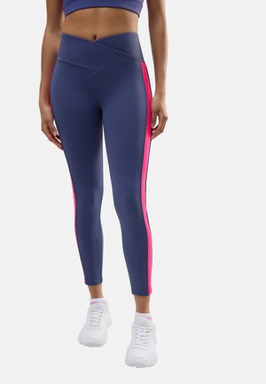 Leggings azul marino con una franja lateral rosa. Material suave y elástico y una cinturilla alta cruzada. Combinados con zapatillas deportivas blancas.