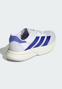 Zapatilla deportiva blanca con detalles morados y un diseño de tres rayas en el lateral. Parte superior de malla ligera y suela acolchada para mayor soporte.