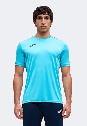Homme portant un t-shirt de sport bleu clair à manches courtes et un pantalon bleu foncé avec des logos assortis de la marque sportive, debout devant un fond uni.