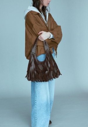 Personne en veste marron et sweat à capuche gris tenant un sac à main en cuir marron foncé à franges, portant un jean bleu clair sur un fond uni.