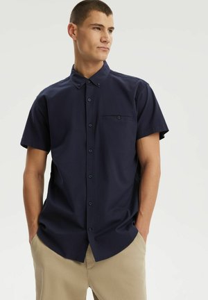 Młody mężczyzna stoi z rękami w kieszeniach, ubrany w granatową zapinaną na guziki krótką koszulę i beżowe spodnie, patrząc na bok.