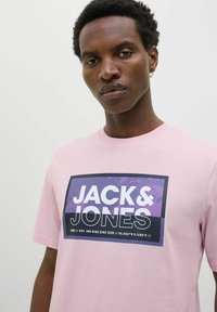 Lyserød T-shirt med korte ærmer, der har et stort sort og lilla grafisk design, som tydeligt viser "JACK & JONES" på forsiden.