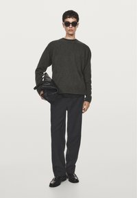 Maglione di lana verde scuro con scollo a girocollo, abbinato a pantaloni scuri. Tiene una borsa in pelle nera. Occhiali da sole neri completano il look.