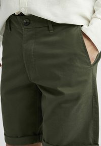 Shorts en coton verts avec une texture lisse, dotés d'une fermeture à bouton, de poches latérales et de bords roulés pour un look soigné.
