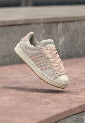 Beżowy zamszowy sneaker z różowymi akcentami, z wiązaniem na sznurowadła, trzema paskami z boku i wzmocnionym gumowym noskiem, na tle teksturowanej powierzchni.