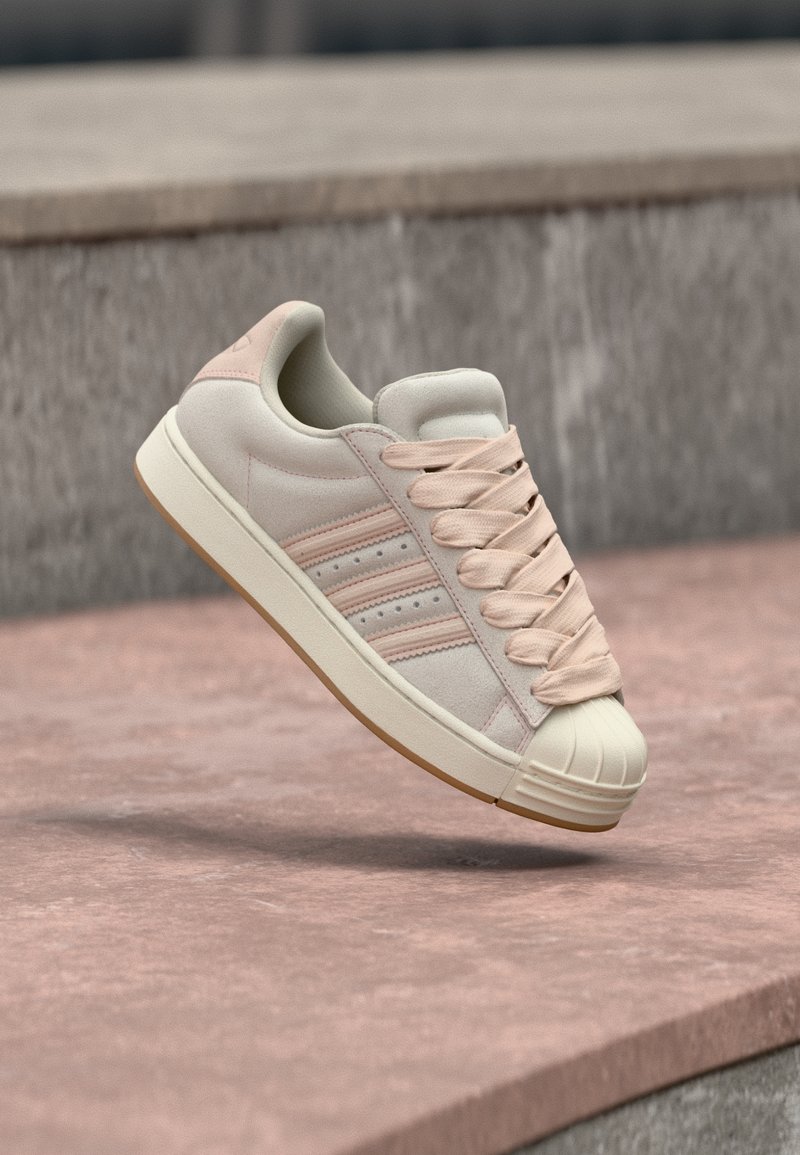 Μπέζ σουέτ αθλητικό παπούτσι με ροζ λεπτομέρειες, διαθέτει κορδόνια, τρεις λωρίδες στο πλάι και ενισχυμένη λαστιχένια μύτη, τοποθετημένο σε υφή επιφάνειας.