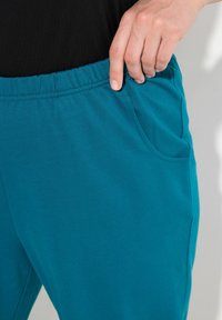 Turkusowe dresy z miękkiego materiału, z elastycznym pasem, bocznymi kieszeniami i gładką teksturą. Ręka oparta na kieszeni.