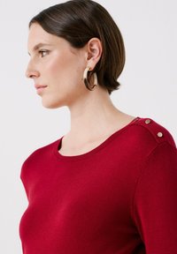 Röd tröja med rundad halsringning, försedd med guldiga accentknappar på axeln, tillverkad av mjukt stickat tyg med en slät yta.