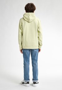 Felpa verde chiaro con cappuccio e cordino, abbinata a jeans blu. La felpa ha una vestibilità comoda e polsini a costine, indossata con sneaker bianche e nere.