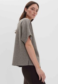 Frau mit glattem braunem Haar, die ein strukturiertes, graues Kurzarmoberteil und dunkle Hose trägt, seitlich blickend mit neutralem Gesichtsausdruck vor schlichtem Hintergrund.