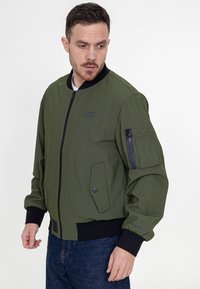 Oliwkowa kurtka bombowca z czarnym ściągaczem, kieszenią na zamek na rękawie, bocznymi kieszeniami oraz gładką fakturą. Posiada nadrukowane logo.